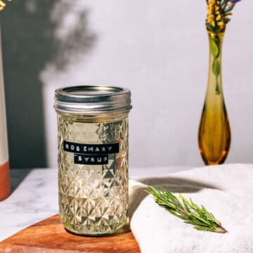 Jar of the syrup on a wood cutting board with a sprig of rosemary on the side.