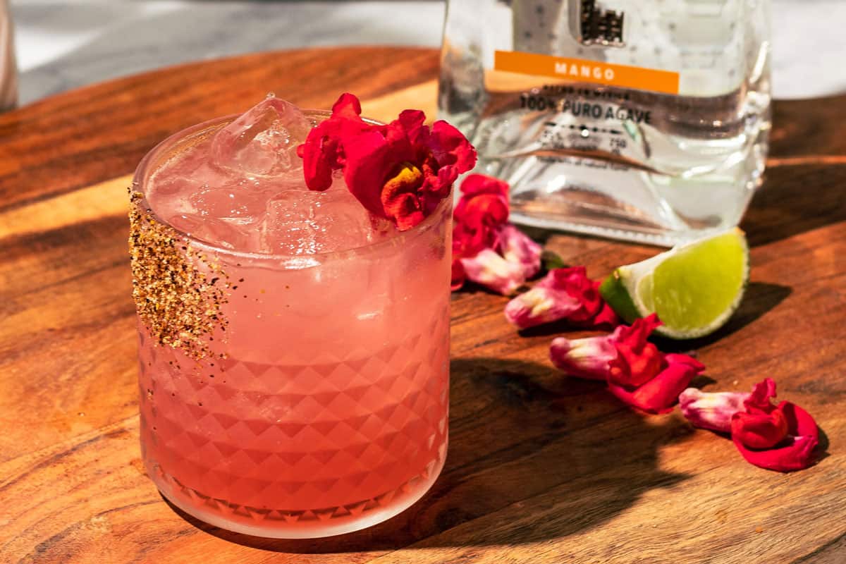 The margarita with a bottle of tequila in the background and scattered flowers on a wood surface.