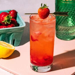 Strawberry gin and tonic on a pink surface with strawberries and lemons in the background.