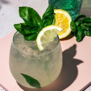 Cocktail sitting on a pink surface with a lemon half and fresh basil leaves in the background.