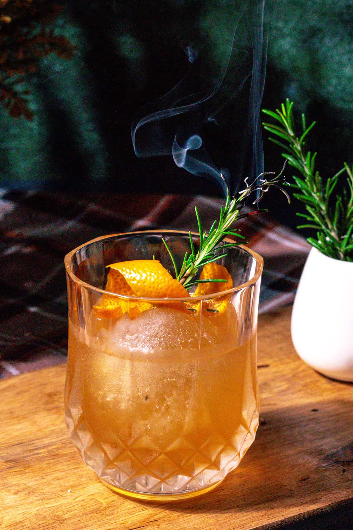 Whisky old fashioned on a wood surface with smoked rosemary garnish.