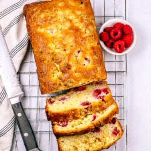 Loaf cake partially sliced on a cooling rack with raspberries on the side.