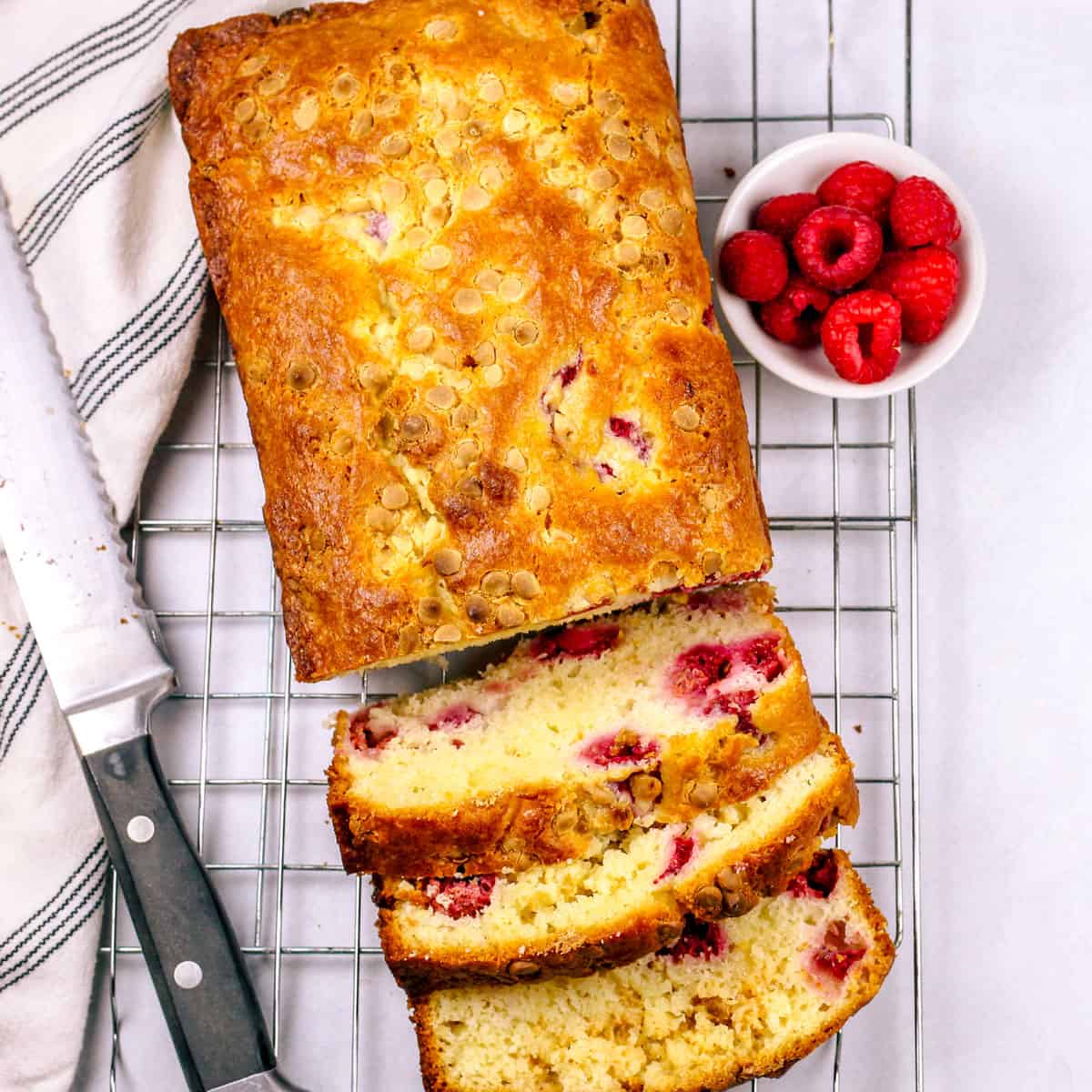 Loaf cake partially sliced on a cooling rack with raspberries on the side.