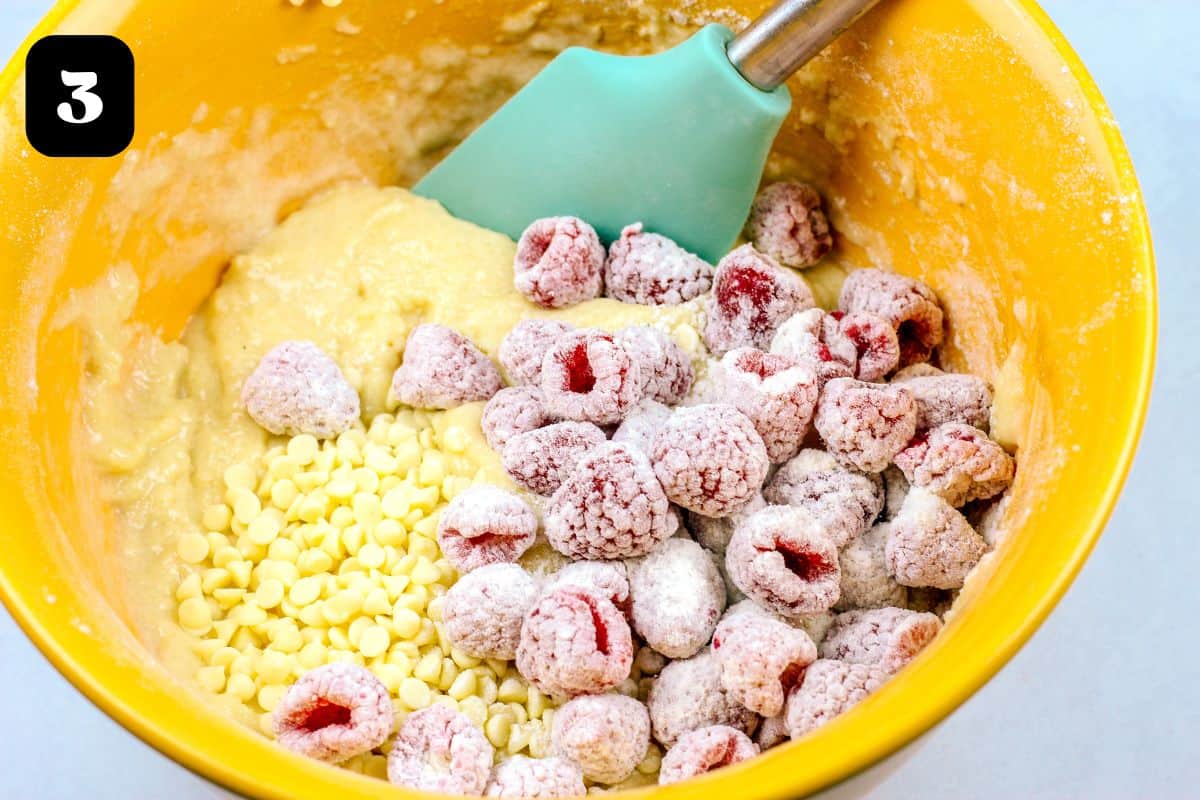 Cake batter with raspberries and white chocolate in a yellow mixing bowl.