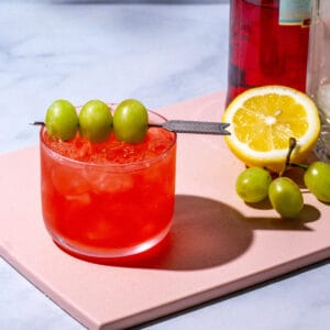 Enzoni cocktail on a pink surface with grapes and a lemon half.
